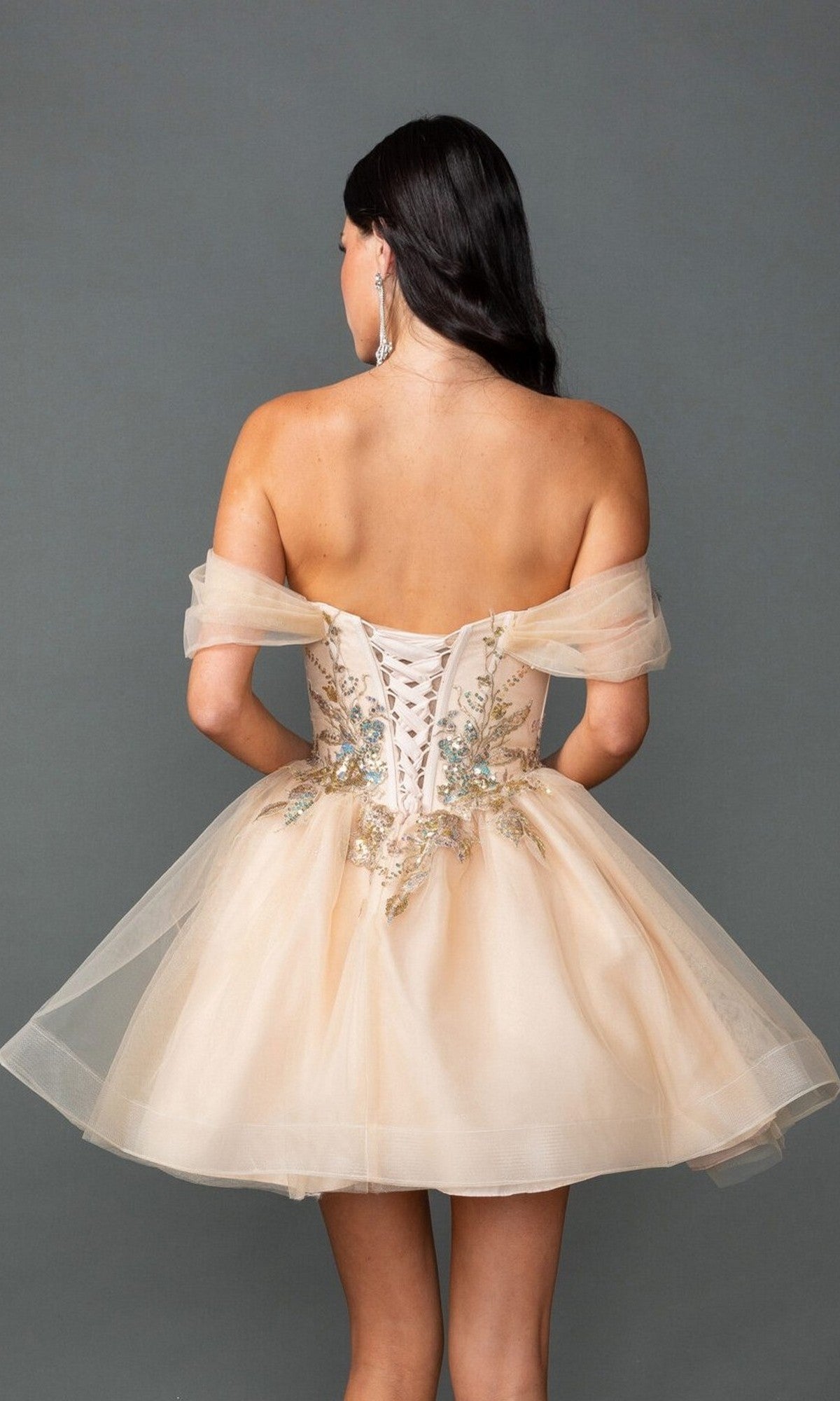 A woman stands facing away, wearing the Dancing Queen 3423 short prom dress in light beige, featuring an off-the-shoulder design, lace-up back, floral embroidery, and an elegant A-line skirt.